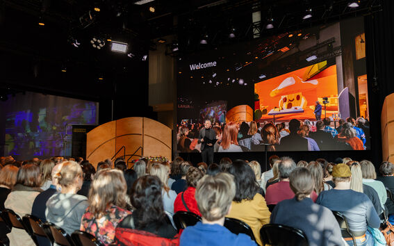 A speaker addresses a large audience in a dark auditorium. A large screen behind shows a vibrant image with the text 'Welcome'.