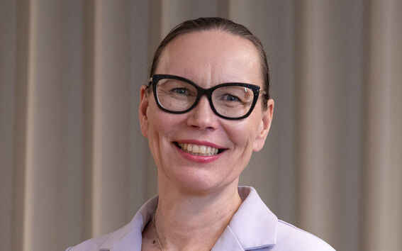 Henkilö, jolla on vaaleanvioletti bleiseri ja sydämenmuotoinen kaulakoru, seisoo beigejen verhojen edessä.