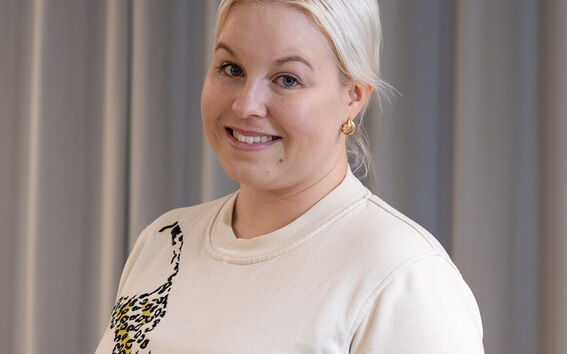 Henkilö, jolla on vaaleat hiukset ja vaalea paita leopardikuvioinnilla, seisoo harmaiden verhojen edessä.