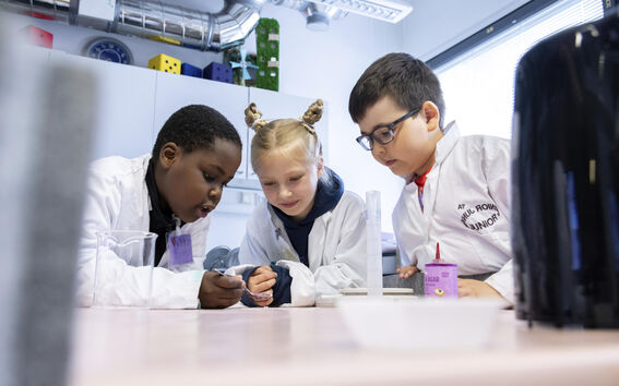 Kolme lasta valkoisissa laboratorio takeissa tekee kokeen luokkahuoneen pöydällä