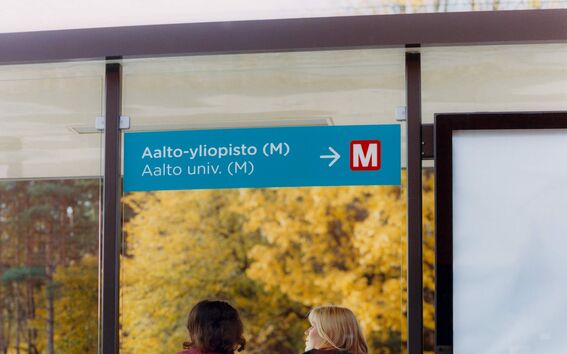 kaksi nuorta istumassa bussipysäkillä, jossa on nuoli Aalto-yliopiston metro-asemalle