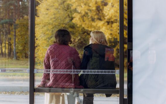 Two people sit at a bus stop near Aalto University, seen from behind with autumn trees in the background