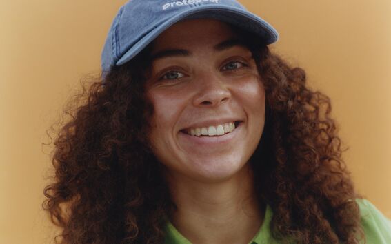 Person wearing a blue cap with text, green shirt, and curly hair against an orange background.