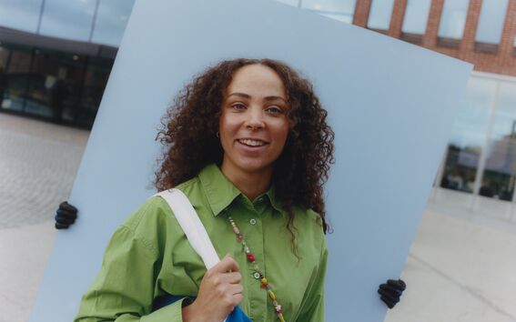 Person in green shirt holding blue bag stands before large grey board held by gloved hands outdoors