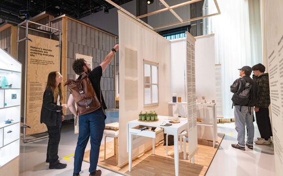 People explore a modern indoor exhibition with wooden walls and white display tables with objects.
