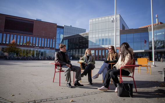 Neljä opiskelijaa juttelee punaisilla ja keltaisilla tuoleilla modernin lasi- ja tiilikampusrakennuksen edessä.