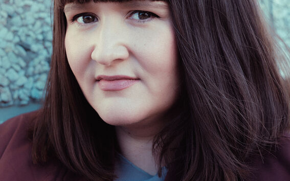 Henkilö, jolla on olkapituinen ruskea tukka, tummapunainen takki ja sininen paita. Taustalla kuvioitu seinä.