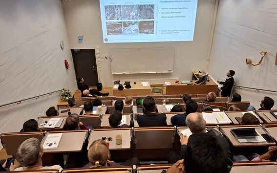 Lecture hall with seated audience watching a man in a dark suit present research slides on a large screen