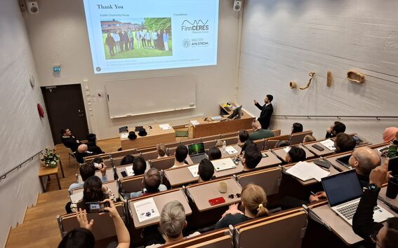 Lecture hall with many people watching a presenter in a suit showing a thank-you slide on a large screen.