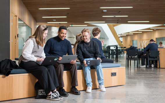 Business school building hall on the ground floor. Three students sitting on a bench and looking at their laptops.