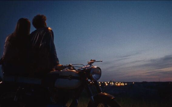Kaksi ihmistä istuu moottoripyörällä illanhämärässä ja katsoo kaukaisia kaupungin valoja.