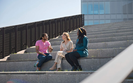 Three students sitting on the amphitheathre steps in front of the business school building.