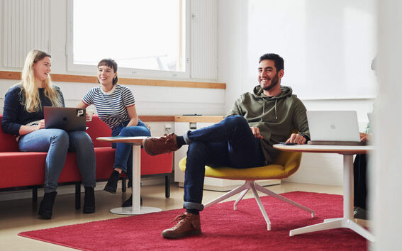 Kaksi ihmistä istuu sohvilla ikkunan edessä keskustellen. Kolmas henkilö istuu etualalla keltaisella nojatuolilla, hän on ristinyt jalkansa ja näyttää nauravan.