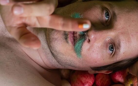 A person lies on a bed of apples, reaching out with their hand. The apples are red, yellow, and green.