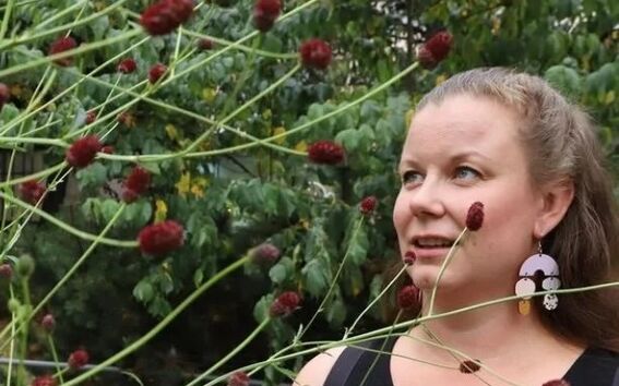 A person in a black sleeveless top is standing behind a bush with red buds. Green foliage is visible in the background.