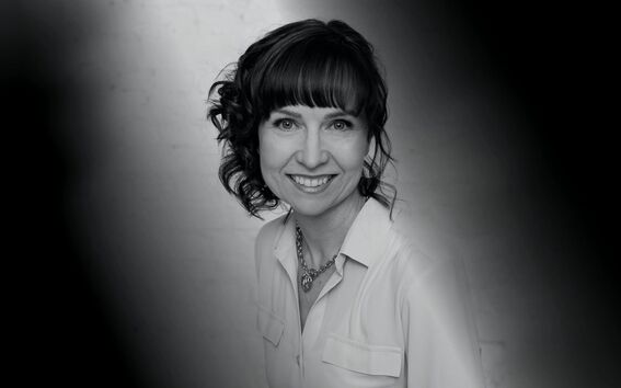 Black and white photo of a person with curly hair, wearing a necklace and a light-coloured blouse with pockets.