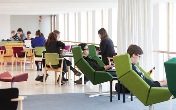 People sitting on colourful chairs at desks in a bright open space, working on laptops or reading.