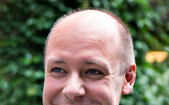 Hymyilevä henkilö, joka käyttää tummaa puvuntakkia ja valkoista paitaa vihreiden lehtien taustalla.