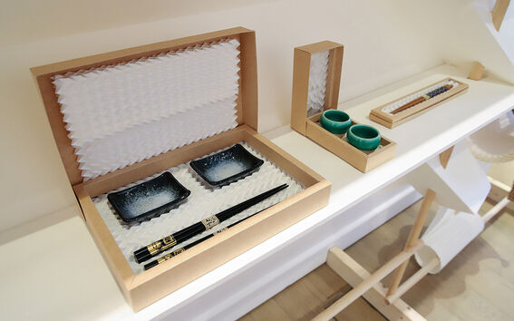 Lahjarasioita valkoisella hyllyllä, joissa syömäpuikot, kaksi mustaa lautasta ja kaksi vihreää kuppia.