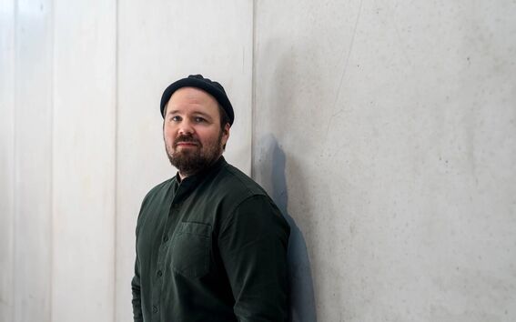A person wearing a dark green shirt and black hat leans against a light grey concrete wall.