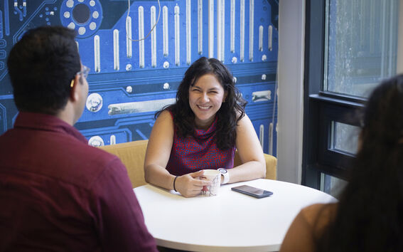 Three people talk at a round table; woman holds a cup, phone nearby, tech wall behind