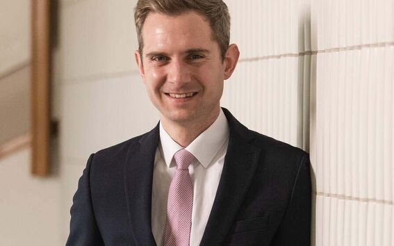 Person in dark suit and pink tie leaning against a tiled wall, hands in pockets