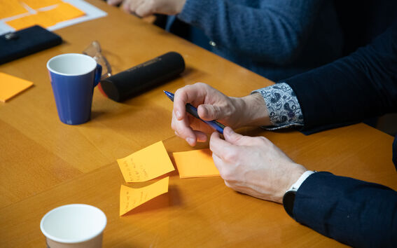 People at a meeting table with notes, a pen, mugs and a speaker on the wooden surface