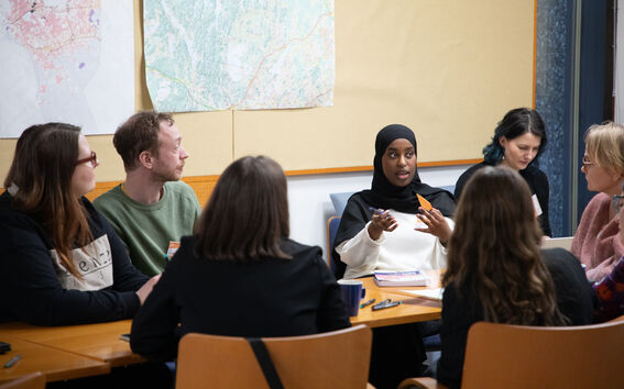 Group of people sit around a wooden table discussing, with large maps pinned on the wall behind them.
