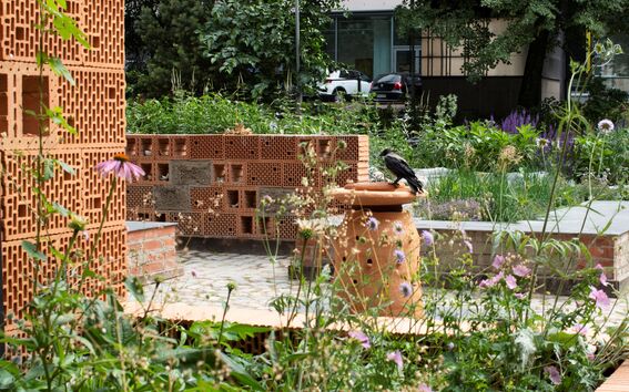 Vehreä puutarha, luonnonkukkia ja tiilirakenteita, lintu istuu saviruukkupylväällä.