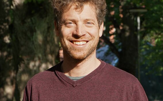 Person with blurred face in a maroon jumper standing in a sunlit park with large trees