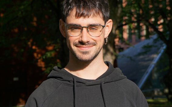Person in black hoodie with white logo standing in a sunny park, trees and building in background