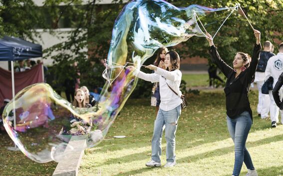 Two people outdoors making a huge rainbow soap bubble on green grass, others watching in the background