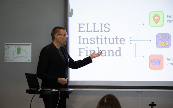 Person in dark suit presenting ELLIS Institute Finland slide with colourful icons in a lecture room