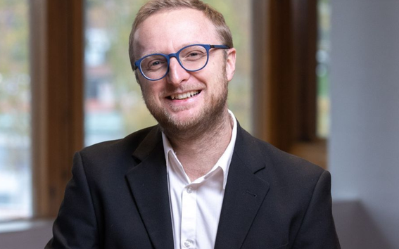 Henkilö mustassa bleiserissä ja valkoisessa paidassa istuu sisällä suurten puukarmisten ikkunoiden äärellä