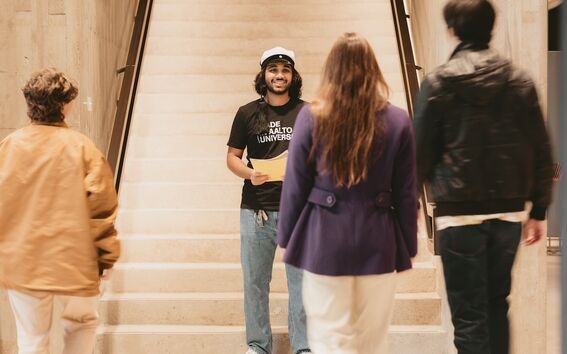 Student in black Aalto University shirt stands on indoor stairs facing three approaching people
