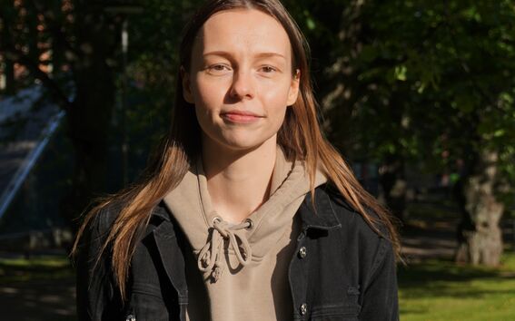 Person in beige hoodie and black jacket standing in a sunny park with green trees