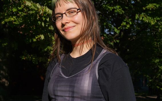 Person with blurred face in dark layered top and light pants, standing on green grass under leafy trees