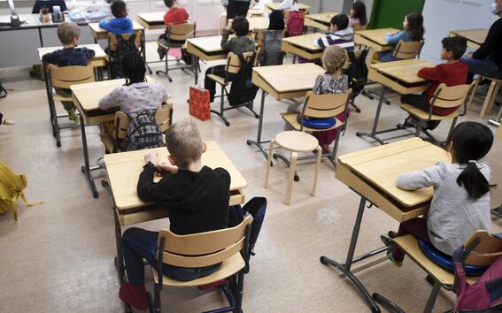 Alakoululaiset istuvat puupulpeteissa valoisassa luokassa ja katsovat kohti opettajaa.