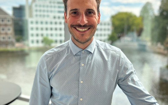 Vaaleansinipaitainen henkilö ikkunan äärellä, taustalla joki ja toimistorakennuksia, kasvot sumentuneet