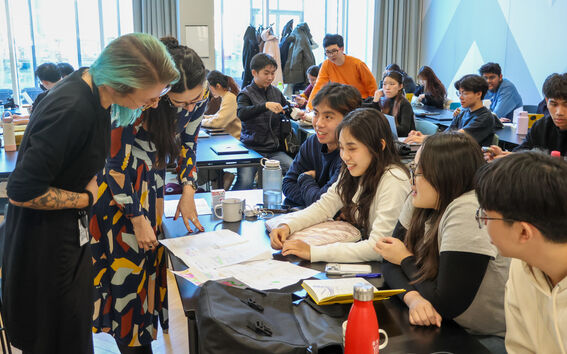 Crowded classroom where two teachers help students gathered around a shared desk with papers and laptops
