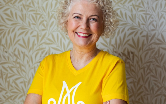 Kiharat vaaleat hiukset omaava henkilö keltaisessa logopaidassa, kädet ristissä, lehtitapetti taustalla