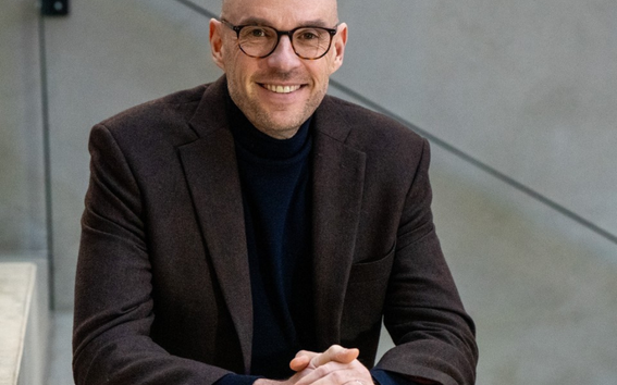 Kalju henkilö tummassa bleiserissä istuu vaalean pöydän ääressä kädet ristissä