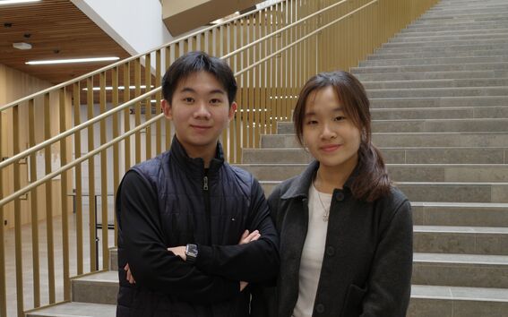 Two people in dark jackets stand by wide indoor stairs with wooden railings and modern lighting