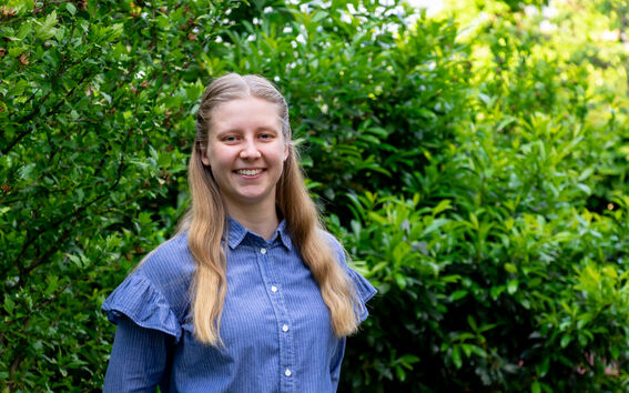Person with long hair in a blue shirt standing in front of dense green bushes