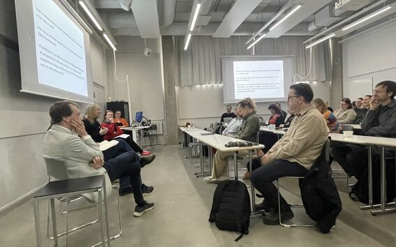 People in a modern classroom listening to a front panel, with slides projected on two white screens.