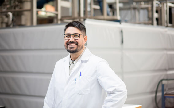 Person in a white lab coat stands in an industrial facility, smiling, blue pen in pocket.