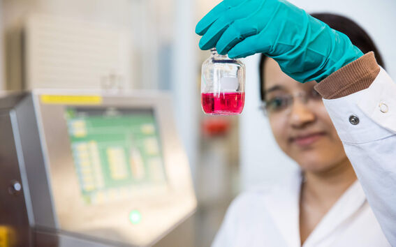 Lab worker holds clear vial of pink liquid near a machine with a green-screen display