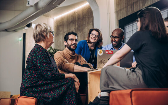 Group of adults sit indoors around a low table, talking and working on laptops in a modern office.
