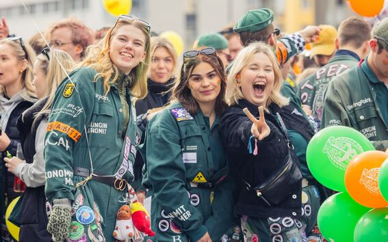 Studenter i gröna overaller på ett livligt utomhusevenemang, ballonger och en gör fredstecken