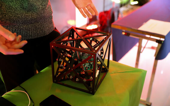 Person presenting a black open cube frame holding electronic circuit boards on a green table